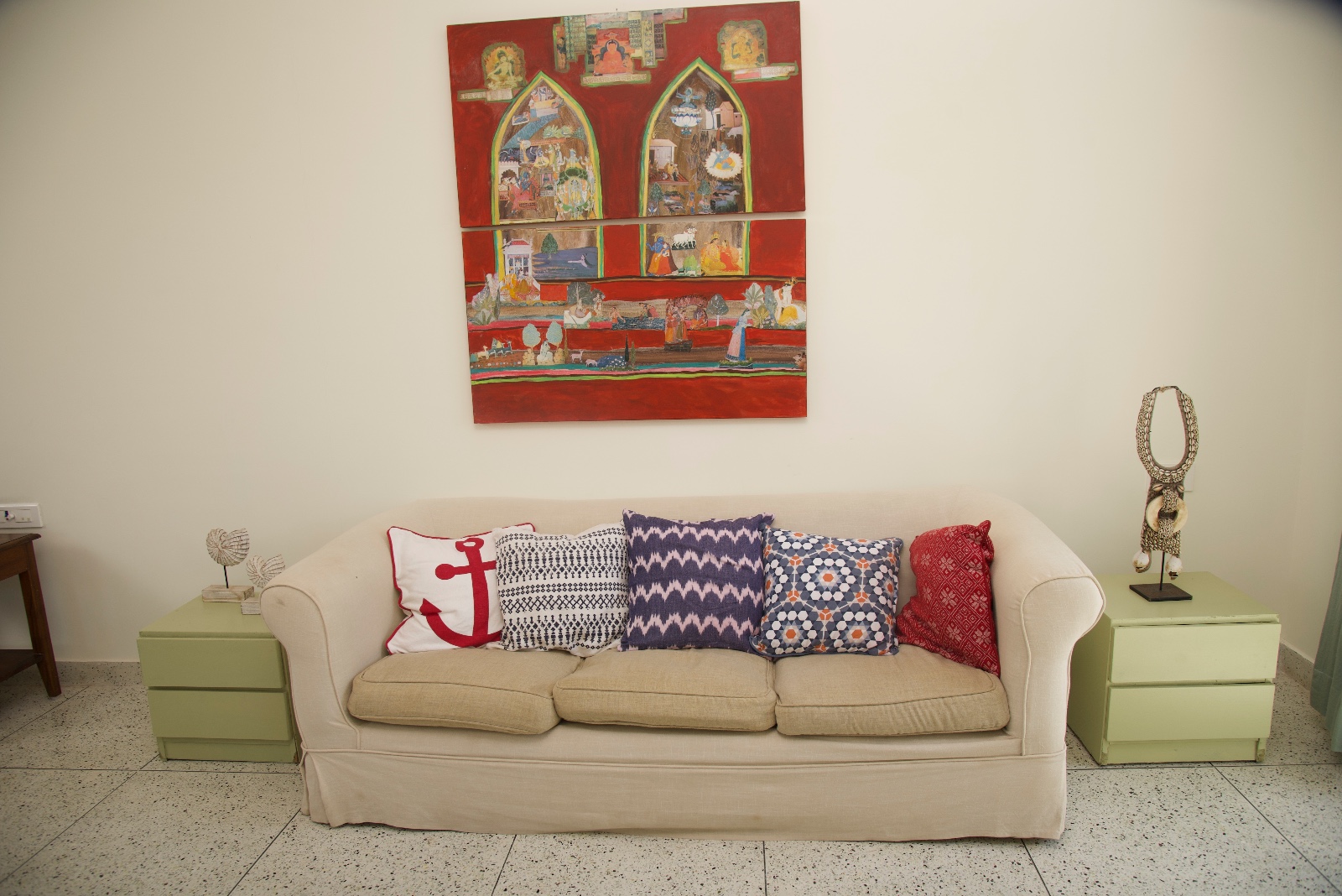 Cream sofa with colourful patterned cushions beneath red artwork and beside pale green tables